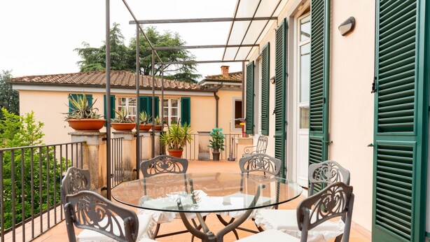Una mesa redonda de vidrio con cuatro sillas, un balcón con barandilla metálica, plantas en macetas y un edificio con contraventanas verdes al fondo.