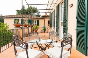 A round glass table with four chairs, a balcony with a metal railing, potted plants, and a building with green shutters in the background. - Apartment 'Lucca Storica' with Private Terrace, Balcony and Wi-Fi (Lucca)