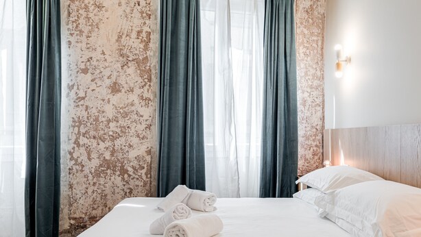 Un lit bien fait, avec des serviettes pliées, une tête de lit en bois et un mur dont la peinture s’écaille.