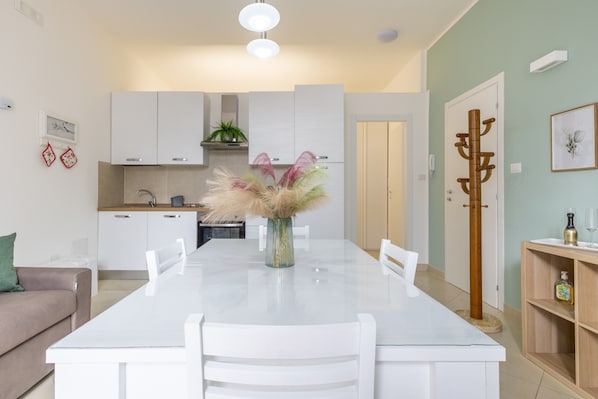 Una cocina moderna con armarios blancos, una mesa de comedor con sillas, un florero con plantas secas y un perchero.