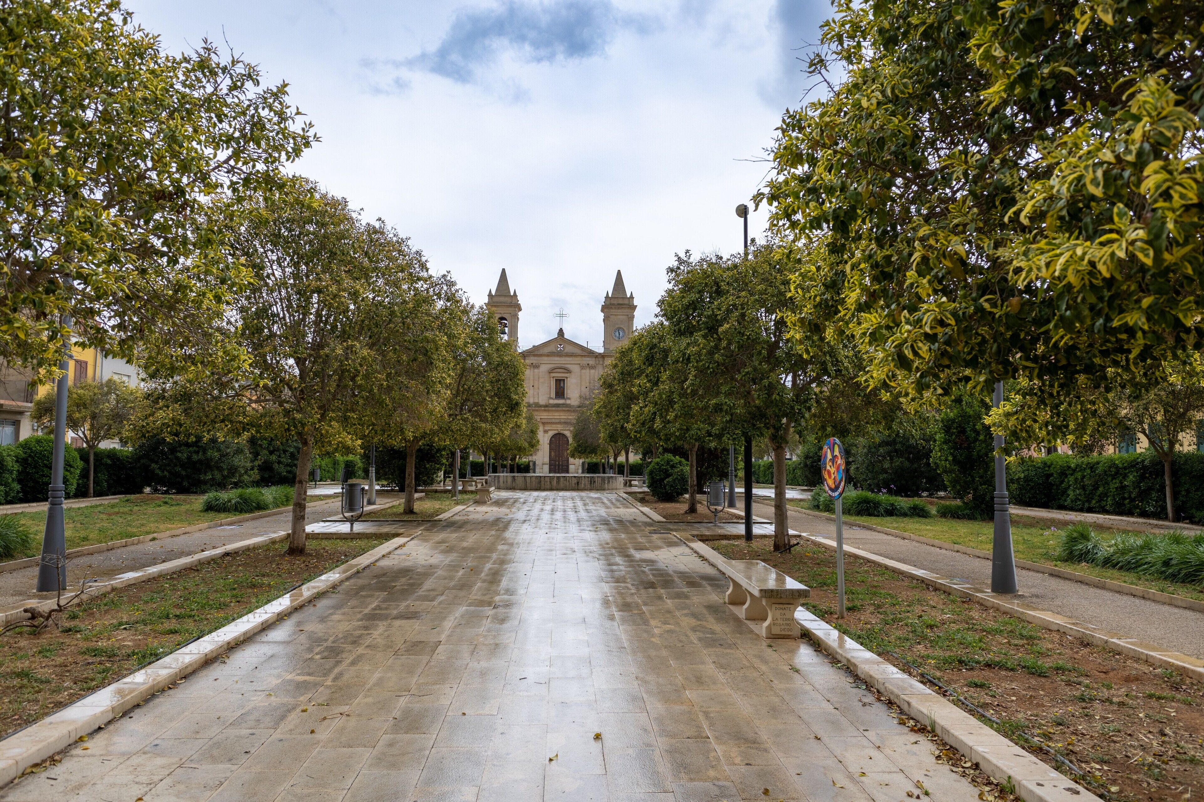 Un chemin pavé mène à un bâtiment doté de deux tours, entouré d’arbres et de bancs.
