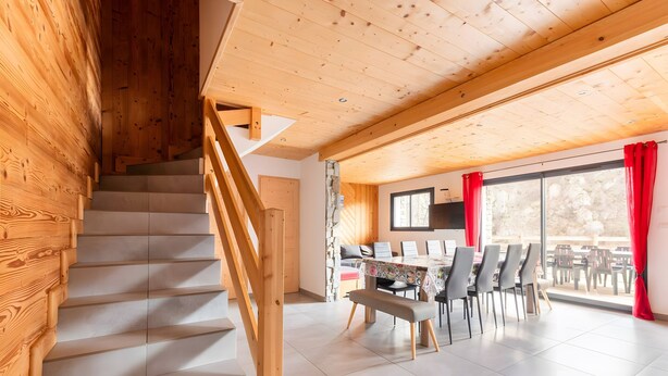 Een houten trap leidt naar een eetruimte met een tafel, stoelen, een bank en uitzicht door grote ramen.