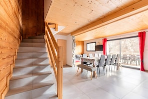 A wooden staircase leading to a dining area with a table and chairs, a bench, and a view through large windows.