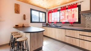 Een keuken met houten kasten, een bar met krukken, een raam met rode gordijnen en een klok aan de muur.