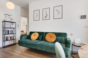 Un canapé vert agrémenté de deux coussins orange, une bibliothèque remplie de livres et trois reproductions botaniques encadrées accrochées au mur.