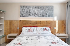 A bedroom with a wooden headboard, a bed with white bedding featuring red and gray patterns, two bedside tables, and a wall-mounted painting of a snowy forest.