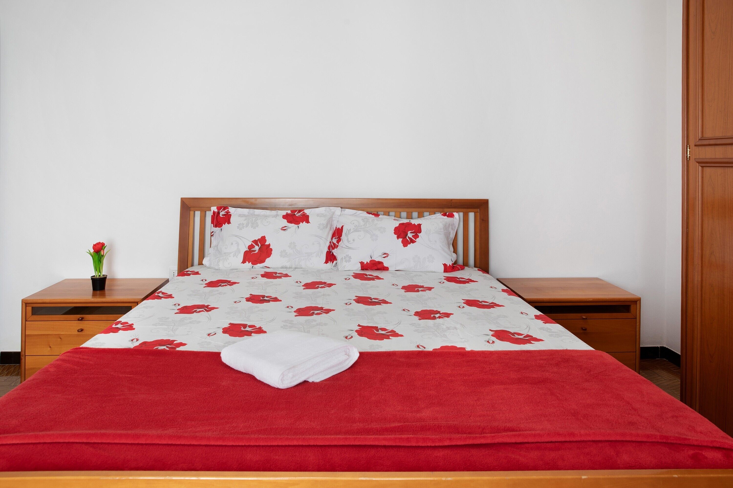 Ein Bett mit einer blumigen Tagesdecke, einer roten Decke und einem weißen Handtuch. Holz Nachttische mit Schubladen und ein kleiner Topfpflanze sind ebenfalls vorhanden.