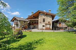A two-story stone building with a wooden balcony, a stone chimney, and a grassy yard.