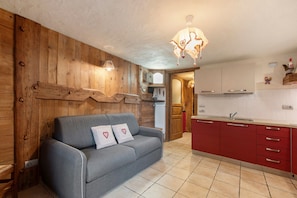 Una habitación con pared de madera, un sofá gris con cojines de estampado de corazones, una cocina roja con armarios blancos y una lámpara colgante.