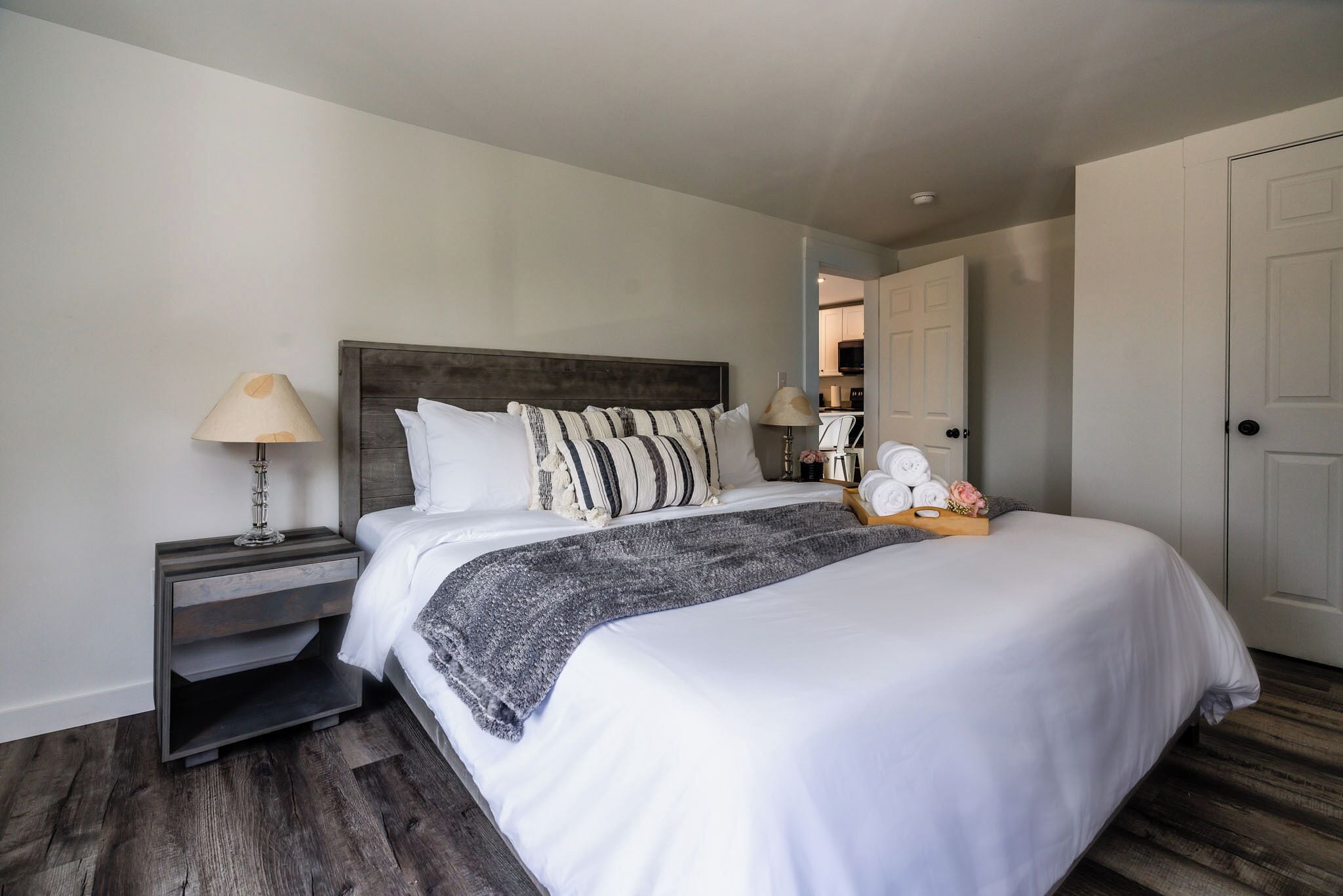 A neatly made bed with white linens and a gray throw blanket, a bedside table with a lamp, and a wooden nightstand.