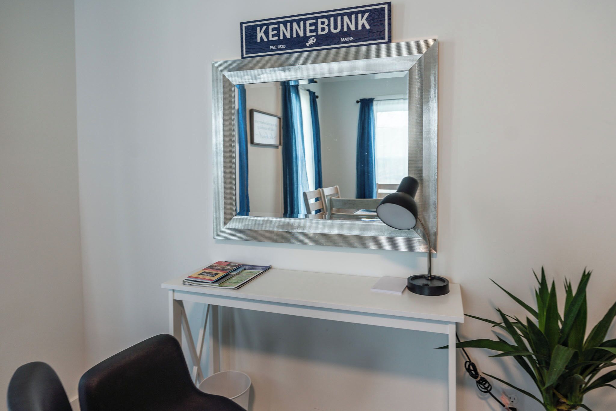 Een kamer met een wit bureau, een stoel, een spiegel, een bord met de tekst 'KENNEBUNK' en een kamerplant.