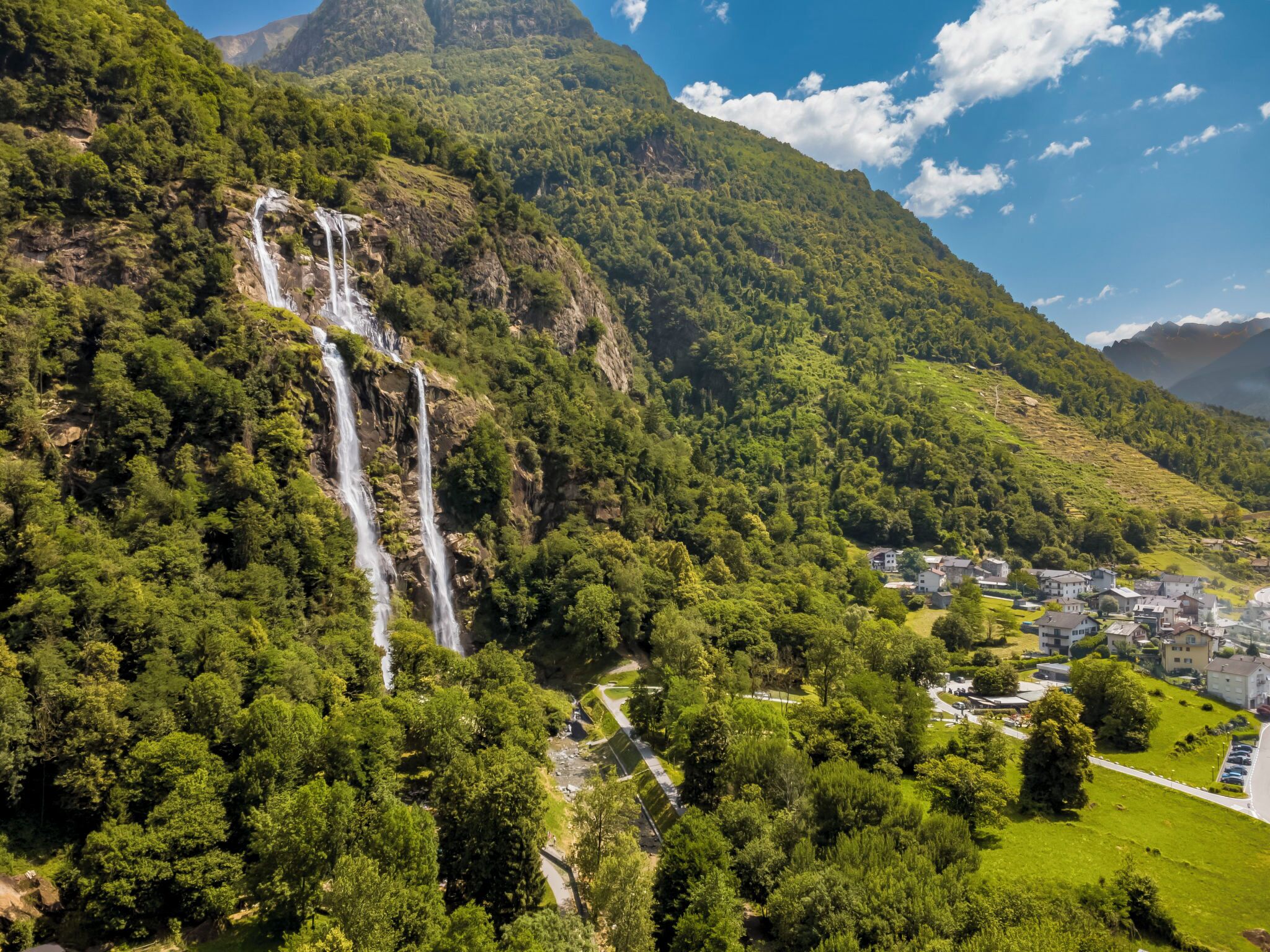 Paisaje montañoso con cascada, abundante vegetación y un pequeño asentamiento con edificios.