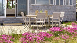 A wooden house with a patio table and chairs, surrounded by pink flowers.