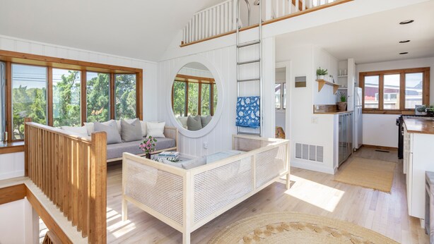 A spacious living area with a high ceiling, a wooden staircase, a round mirror, and a kitchen with white cabinets.