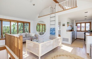A spacious living area with a high ceiling, a wooden staircase, a round mirror, and a kitchen with white cabinets. - Peaceful beach  bungalow on Gerard drive East Hampton Springs (East Hampton)