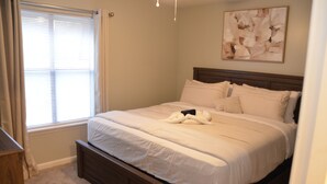 A bedroom with a large bed, a wooden headboard, a window with blinds, and a painting on the wall.