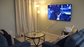 A living room with a TV mounted on the wall, a glass coffee table, and two blue sofas.