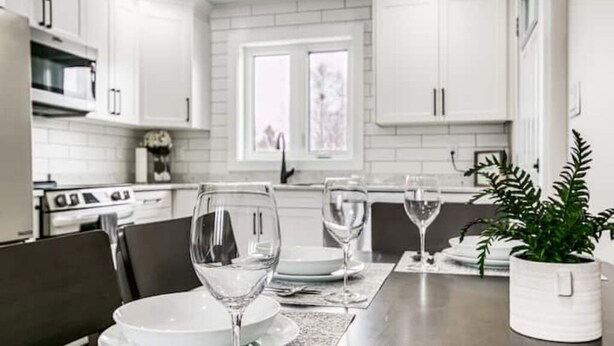 Uma cozinha moderna com armários brancos, uma mesa de jantar posta com pratos e copos de vinho, e um vaso com planta na mesa.