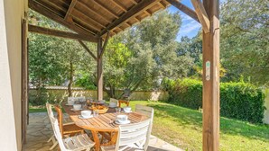 An outdoor patio with a wooden table and white chairs, surrounded by greenery and a stone floor. - Les 2 plages by Interhome (Vaux Sur Mer)
