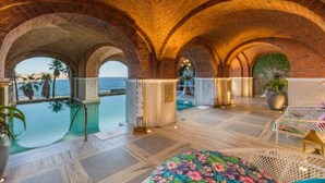 Un espace piscine intérieur luxueux, doté de voûtes en briques, offrant une vue sur la mer et comprenant un coin salon avec un canapé à motifs floraux.