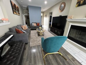 Een woonkamer met een grijze bank, een blauwgroene fauteuil, een zwart piano, een televisie en een open haard.