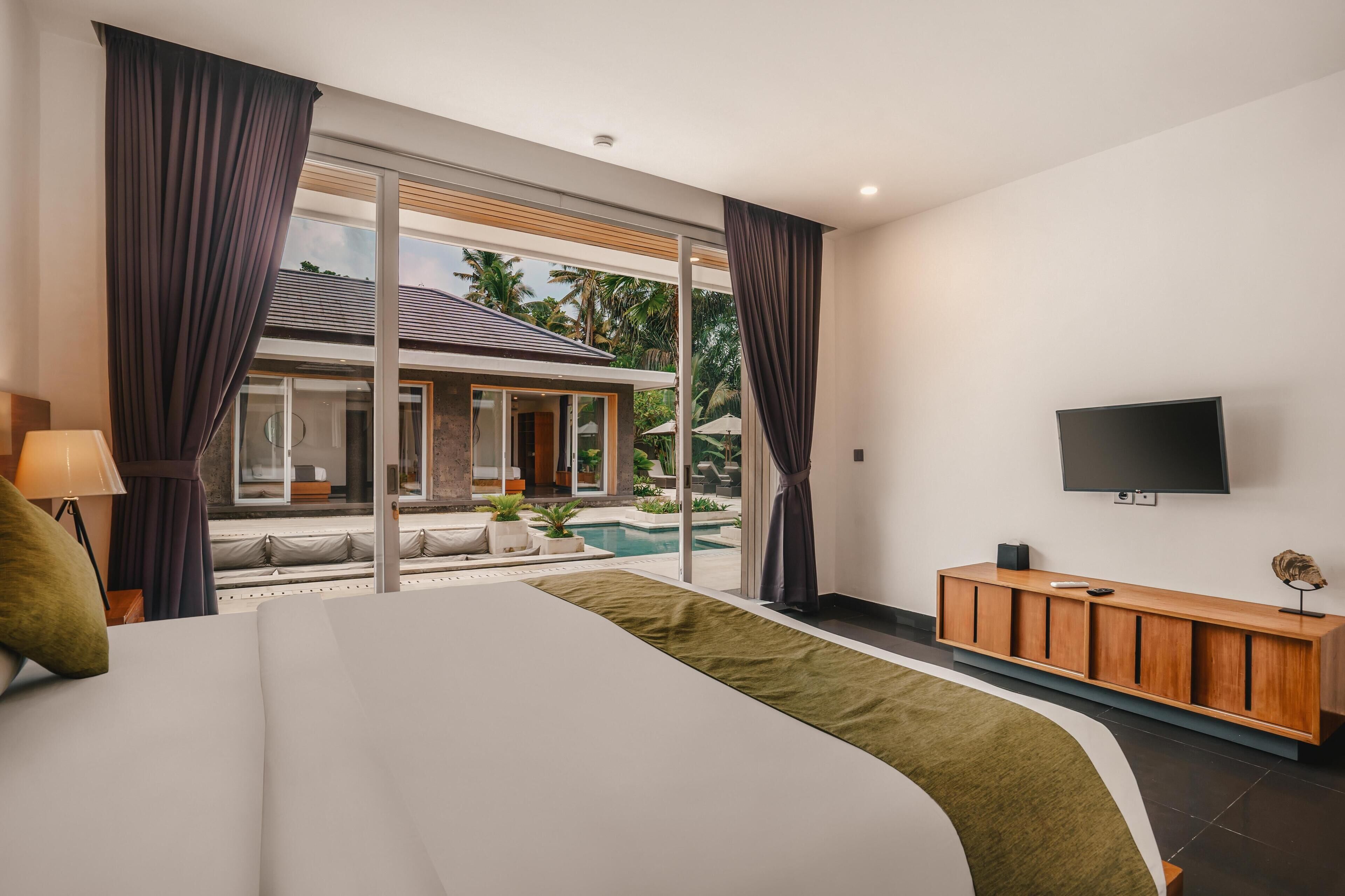 A modern hotel room with a large bed, a TV, a wooden console, and a view of a swimming pool and outdoor area.