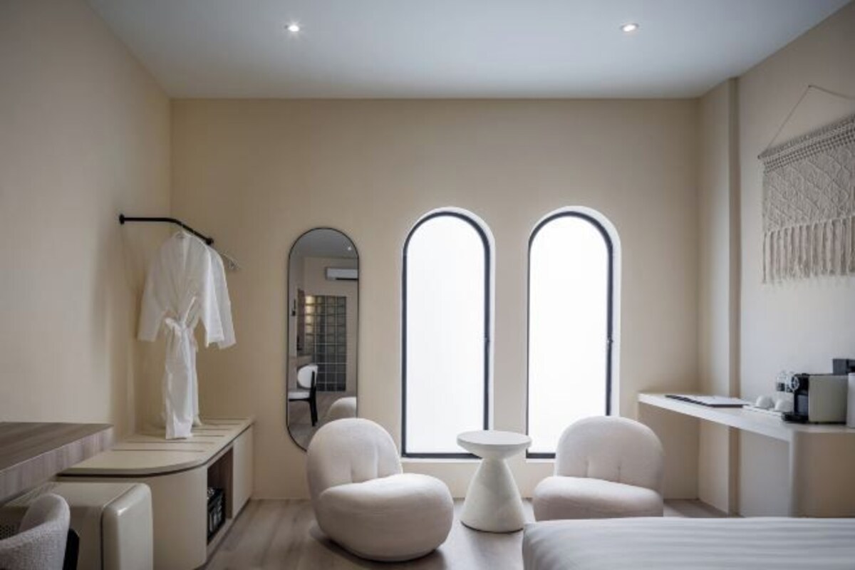 A modern hotel room with a bed, two armchairs, a small table, a desk with a chair, a mirror, and two large windows.
