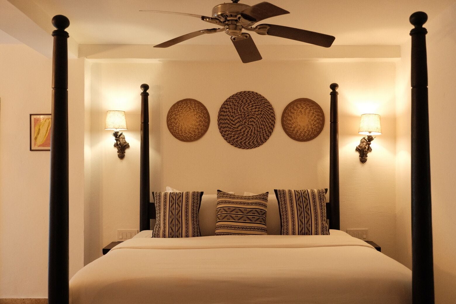 A four-poster bed with patterned pillows, wall sconces, and decorative wall art.