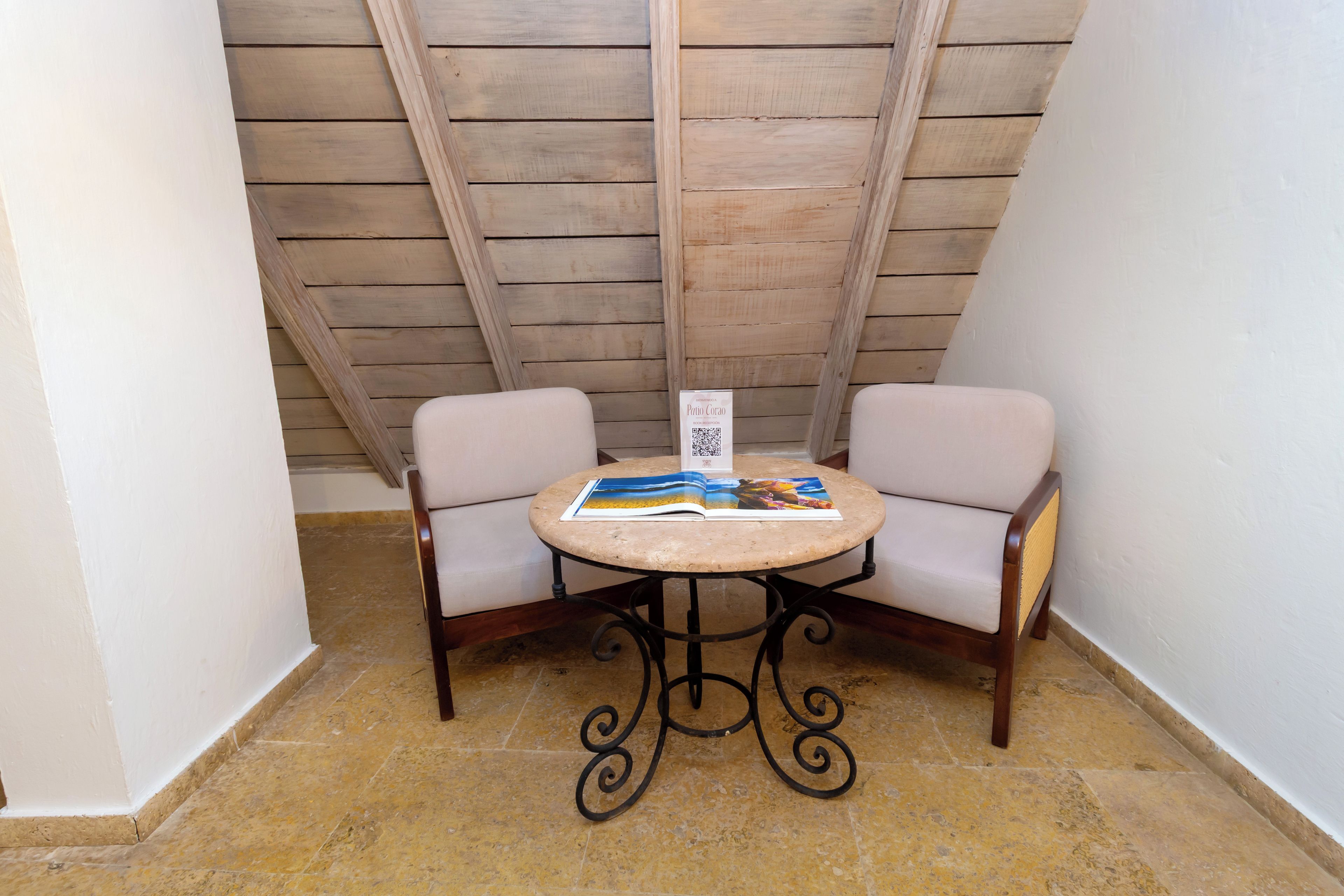 A small, round table with a QR code sign in the center, flanked by two chairs with a tiled floor and wooden slats on the ceiling.