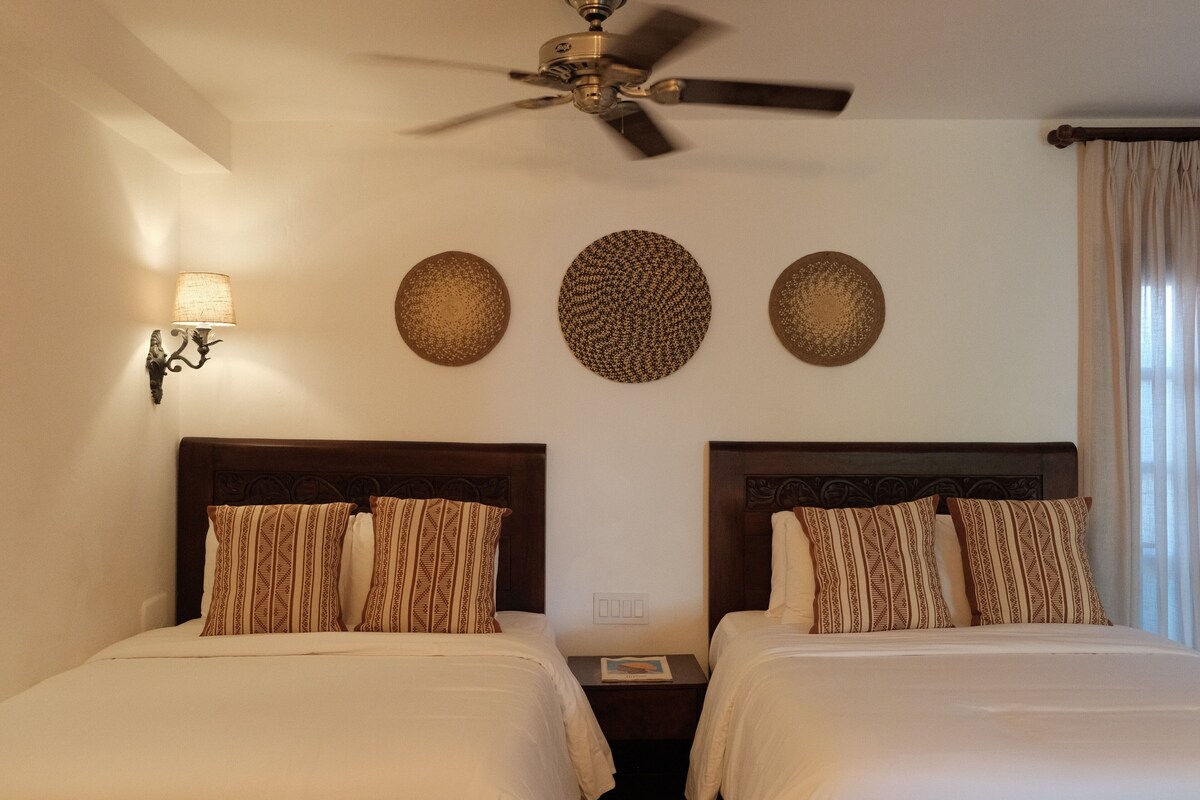 Quarto de hotel com duas camas, um ventilador de teto, uma luminária na parede e três decorações de parede texturizadas.