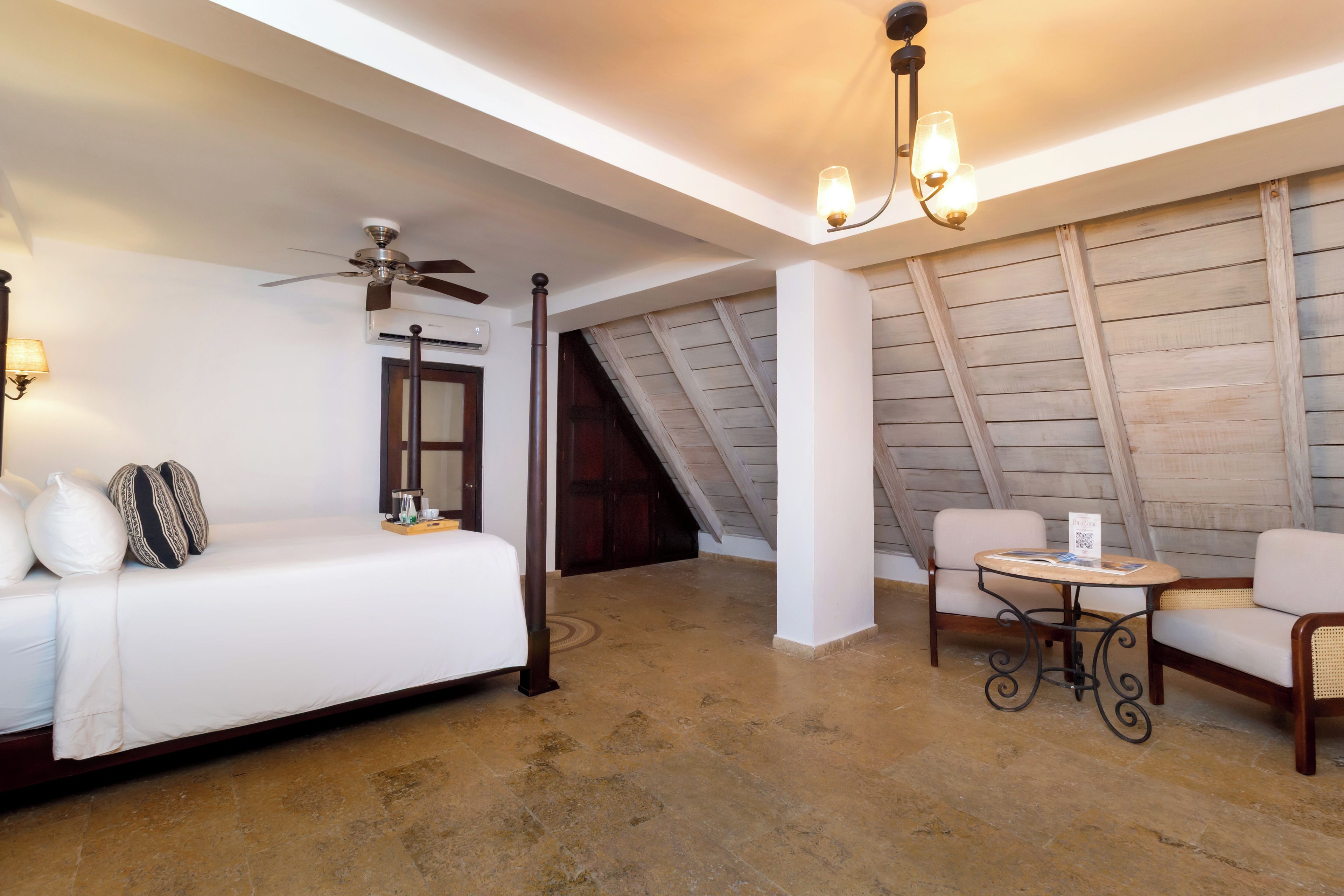 Quarto de hotel com cama, ventoinha no teto, mesa e cadeiras, e vista para o exterior.