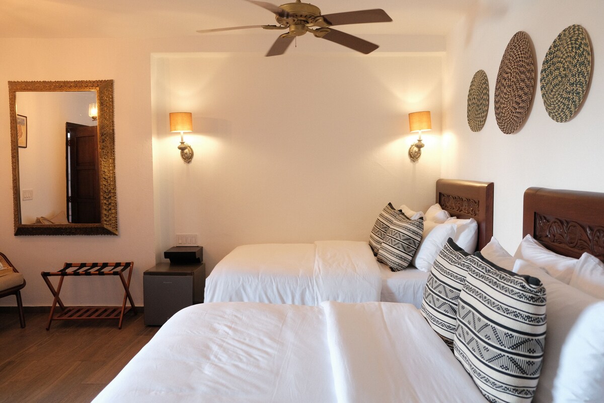 A hotel room with two beds, a ceiling fan, wall-mounted lamps, a small table, and a chair.