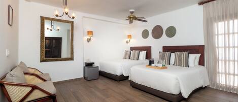 A hotel room with two beds, a sitting area with a chair, a ceiling fan, wall-mounted lamps, and a large mirror.