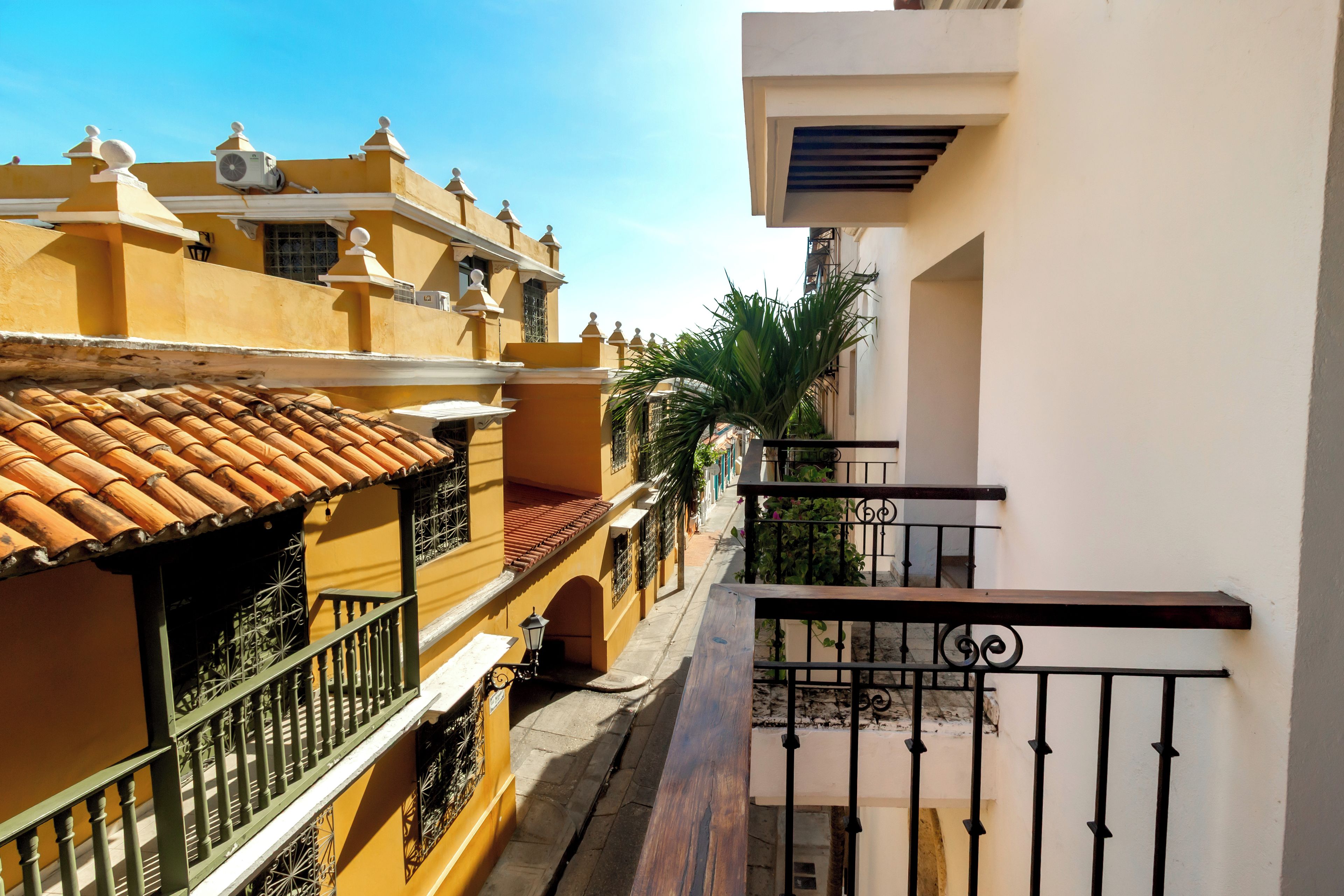 A narrow street with buildings on either side, featuring balconies with metal railings and tiled roofs.