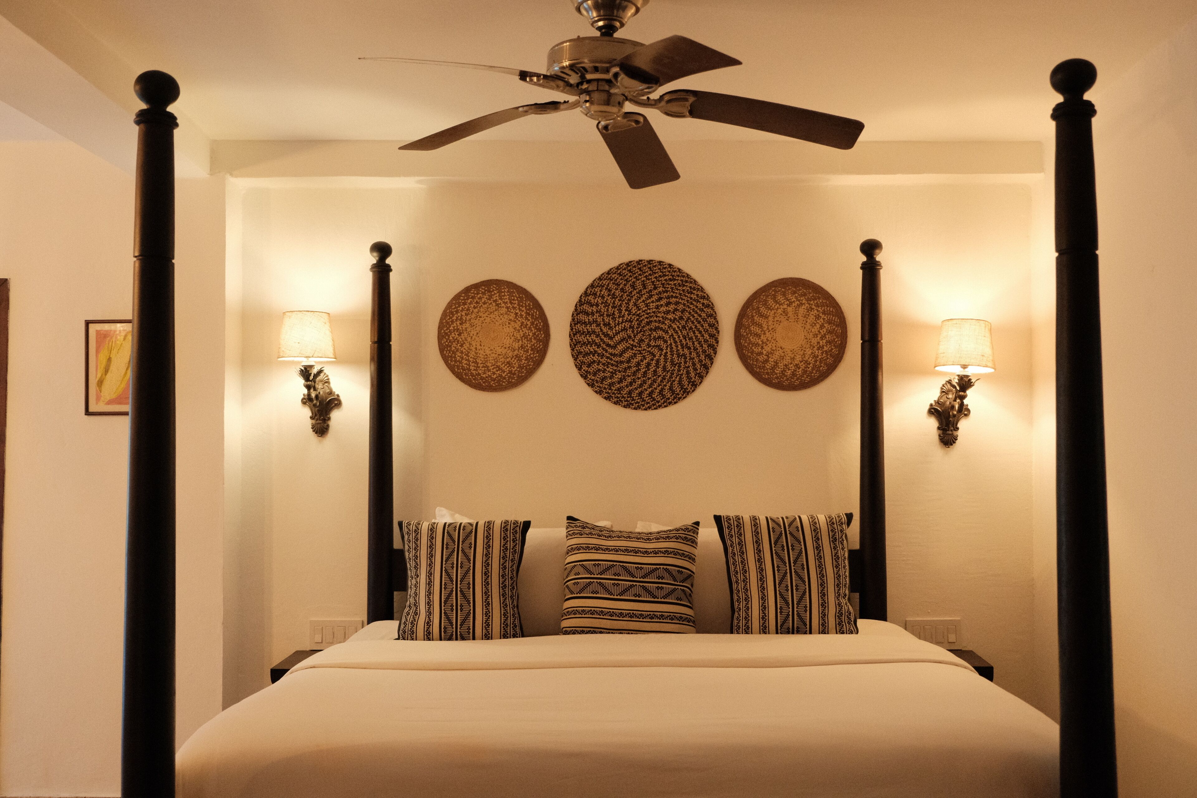 A four-poster bed with a ceiling fan, wall-mounted lamps, and patterned wall decor.