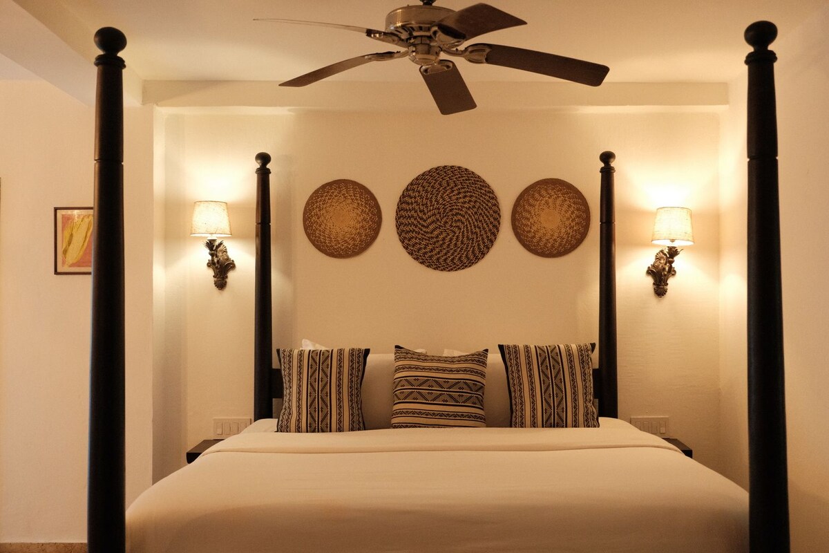 A four-poster bed with patterned pillows, wall sconces, and decorative wall art.