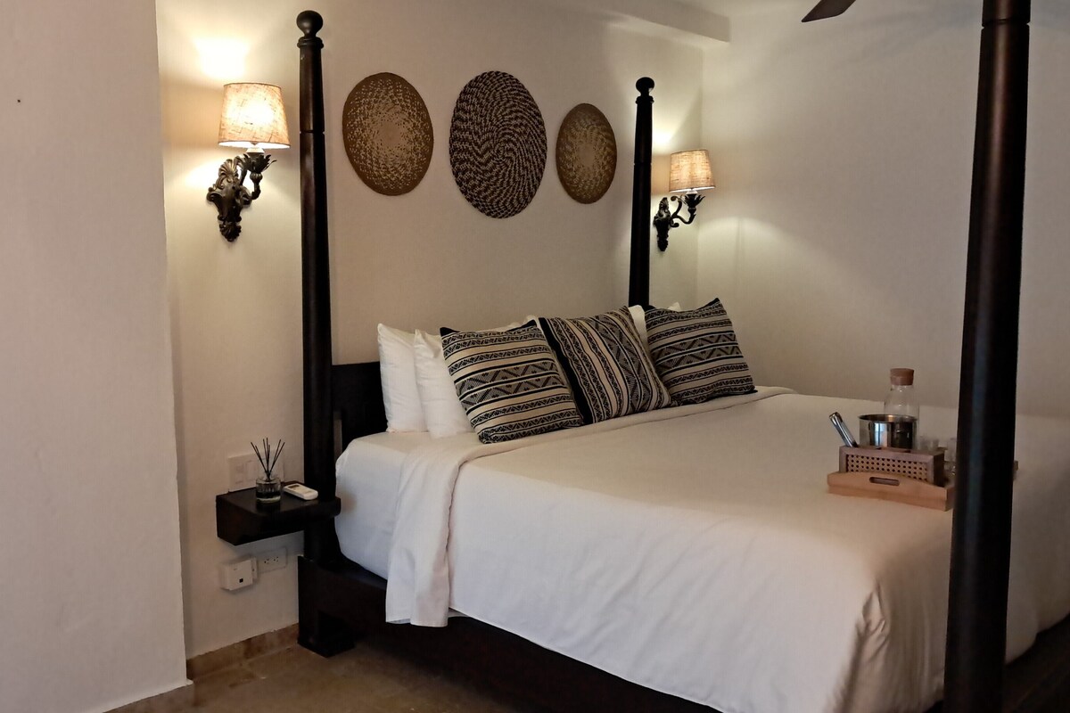 Uma cama bem arrumada com lençóis brancos e almofadas estampadas, uma mesa de cabeceira com abajur, e arandelas na parede.