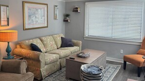 Una sala de estar con sofá, reposapiés, mesa de centro y una ventana con persianas.
