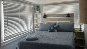 Une chambre à coucher avec un lit, une table de chevet, une lampe, une étagère avec une plante et une fenêtre avec des stores.