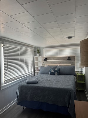 A bedroom with a bed, a bedside table, a lamp, a shelf with a plant, and a window with blinds. - Whole house in Carolina Beach. Walk to beach or bike trail. (Carolina Beach)