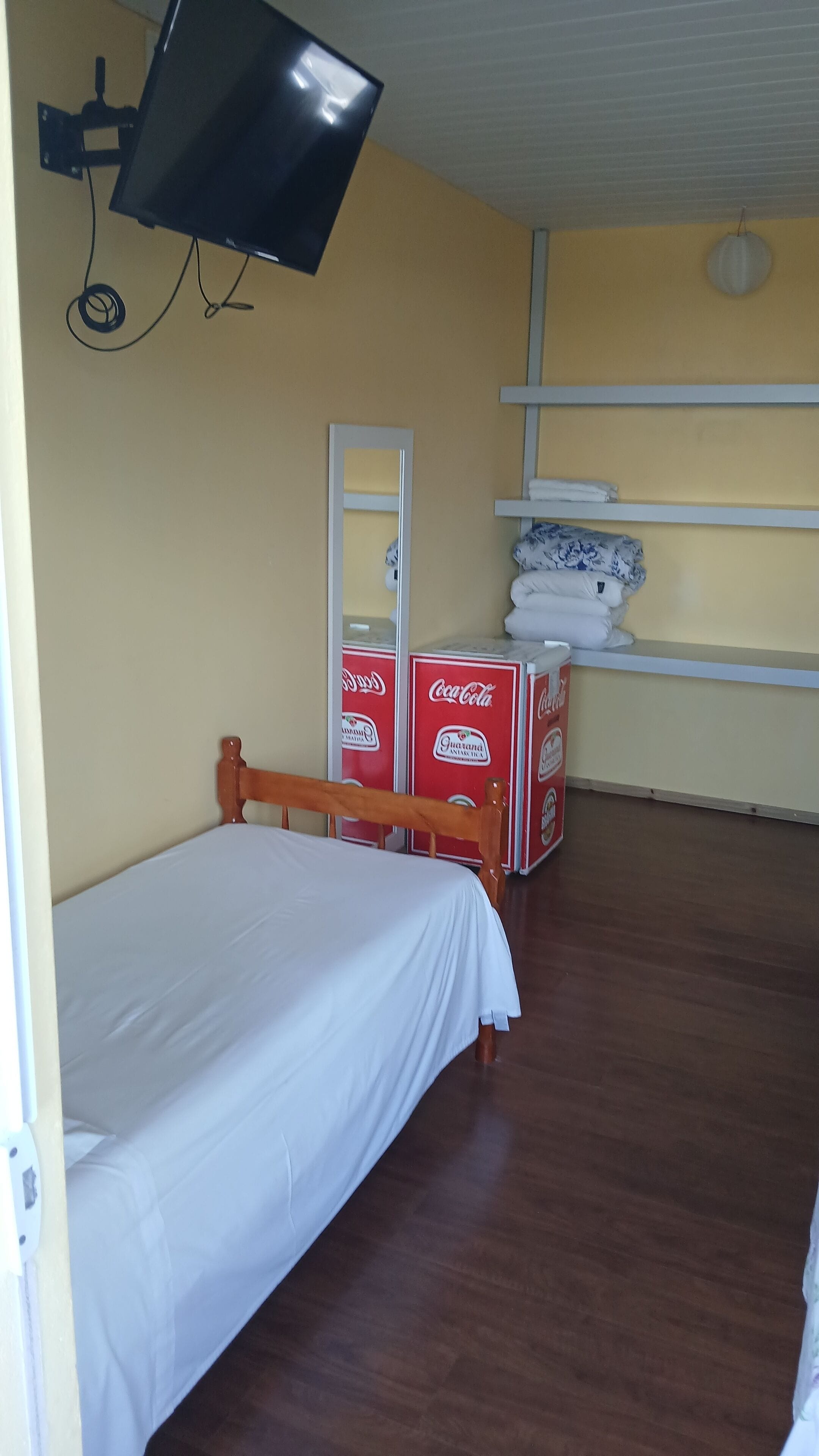 A room with a single bed, a mounted television, a mirror, and stacked Coca-Cola crates.
