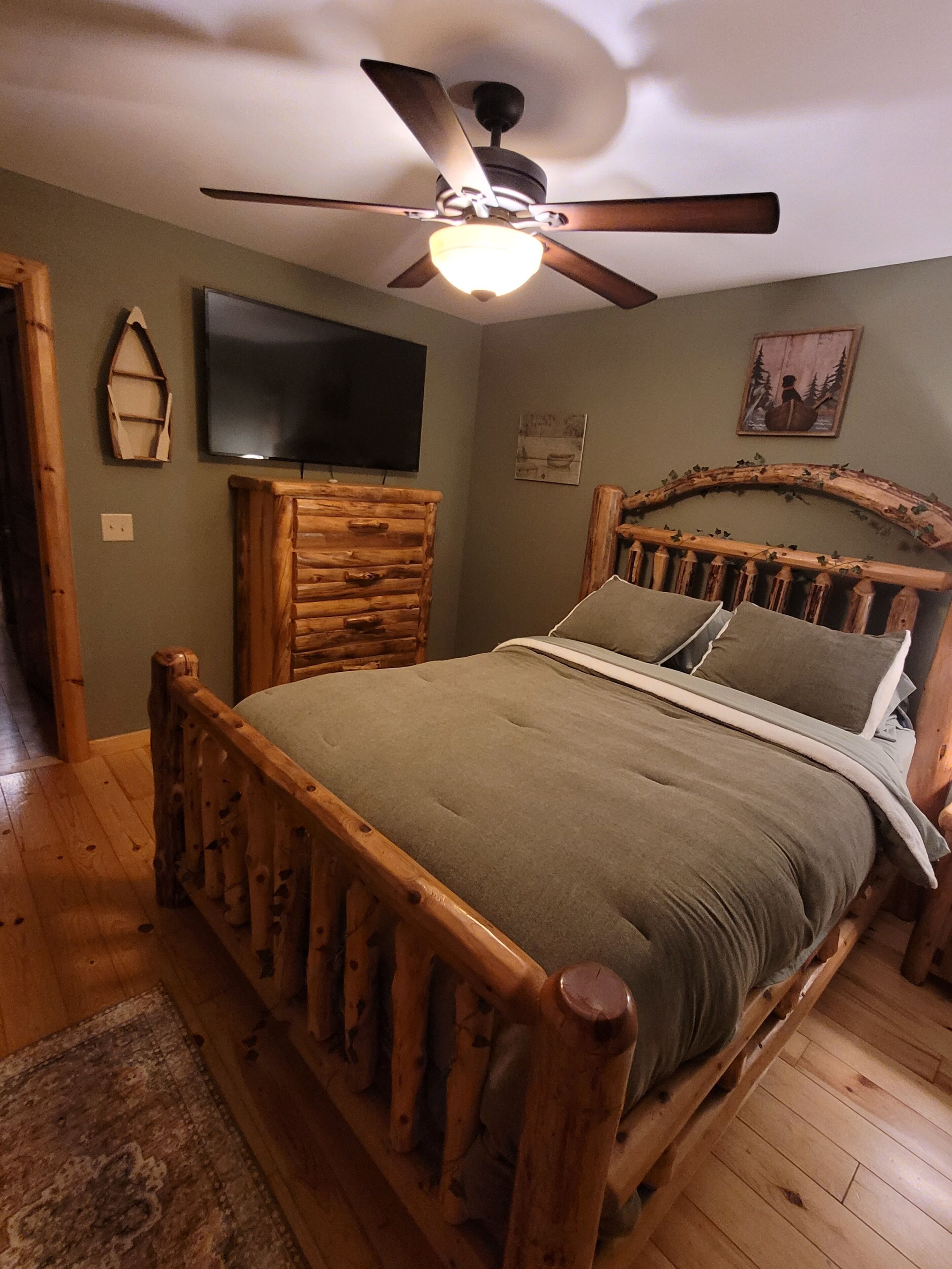 Un lit en bois avec une tête de lit, une commode en bois, un ventilateur de plafond, un téléviseur fixé au mur et un tableau.