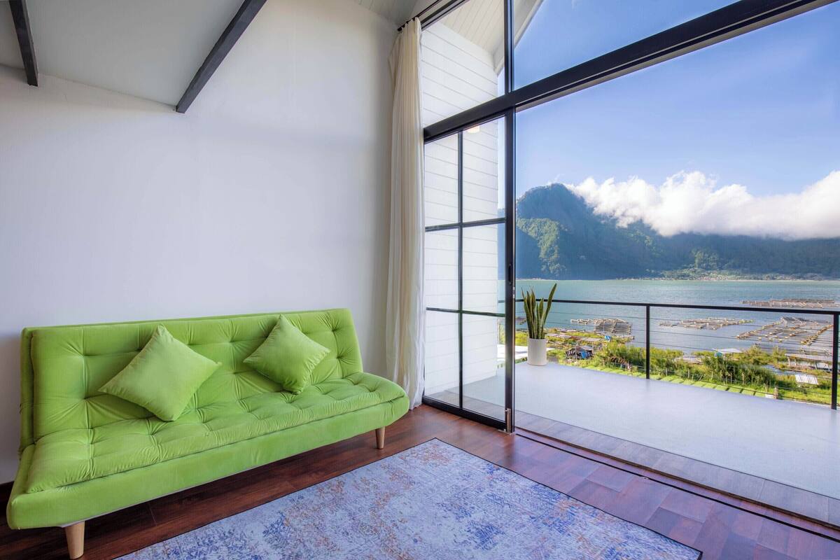 Un salon moderne avec un canapé vert, une grande fenêtre donnant sur un paysage montagneux et une étendue d’eau.
