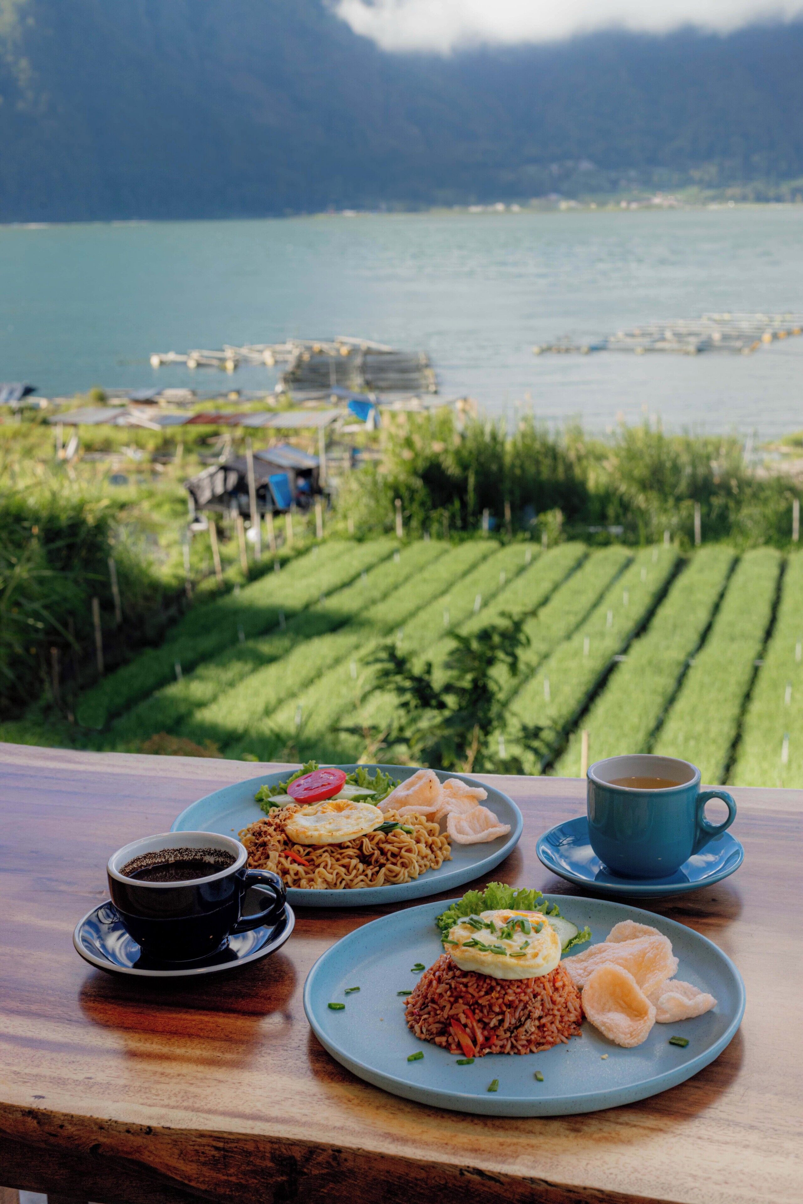 Une table avec deux assiettes de nourriture et une tasse de café, donnant sur une étendue d’eau et un paysage verdoyant.