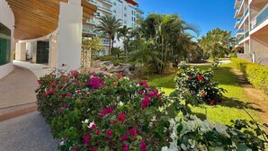 A hotel courtyard with a modern building, palm trees, and a well-maintained garden with pink and white flowers.