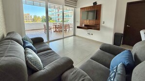 A modern living room with a grey sofa set, a flat-screen TV mounted on the wall, and a sliding glass door leading to an outdoor patio area.