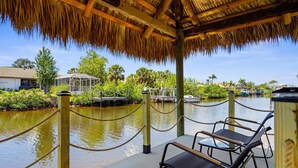 A covered area with a thatched roof, wooden posts, and a rope barrier. There are two black lounge chairs and a small table. - Charming Paradise w/ Beautiful Pool and waterfront Tiki Hut + Backyard Boat Ramp (Port Charlotte)