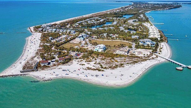 An aerial view of a coastal resort with a long sandy beach, clear blue waters, and a pier extending into the sea. - Charming Paradise w/ Beautiful Pool and waterfront Tiki Hut + Backyard Boat Ramp (Port Charlotte)