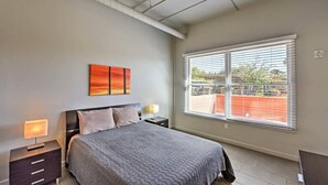 Une chambre d’hôtel moderne avec un grand lit, une table de chevet avec une lampe, une fenêtre avec des stores et un tableau au mur.