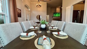 A dining room with a long table set for six, upholstered chairs, a chandelier, and a television in the background. - New House/with Pool/Hot Tub/Putting Green/Close to Universal Studios/Theme Rooms (Frisco)